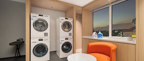 A laundry room with two front-loading washing machines, a small table, an orange chair, and a window with a view of the cityscape.