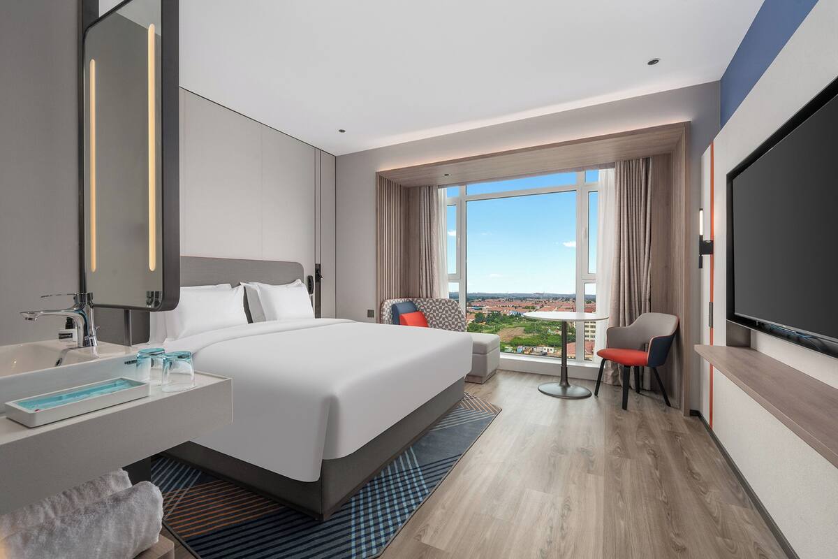 Ein modernes Hotelzimmer mit einem großen Bett, einem Badezimmer mit Waschbecken und Spiegel, einem Fernseher und Blick auf die Stadt.