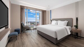A modern hotel room with a large bed, a seating area with a chair and ottoman, a small table, and a large window offering a city view. - Holiday Inn Express Qingdao Hongdao Station by IHG (Qingdao)