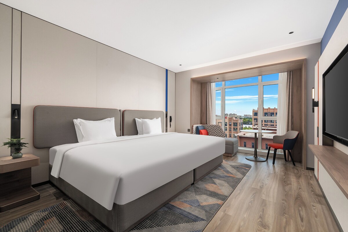 A modern hotel room with a large bed, a desk, and a view of the city.