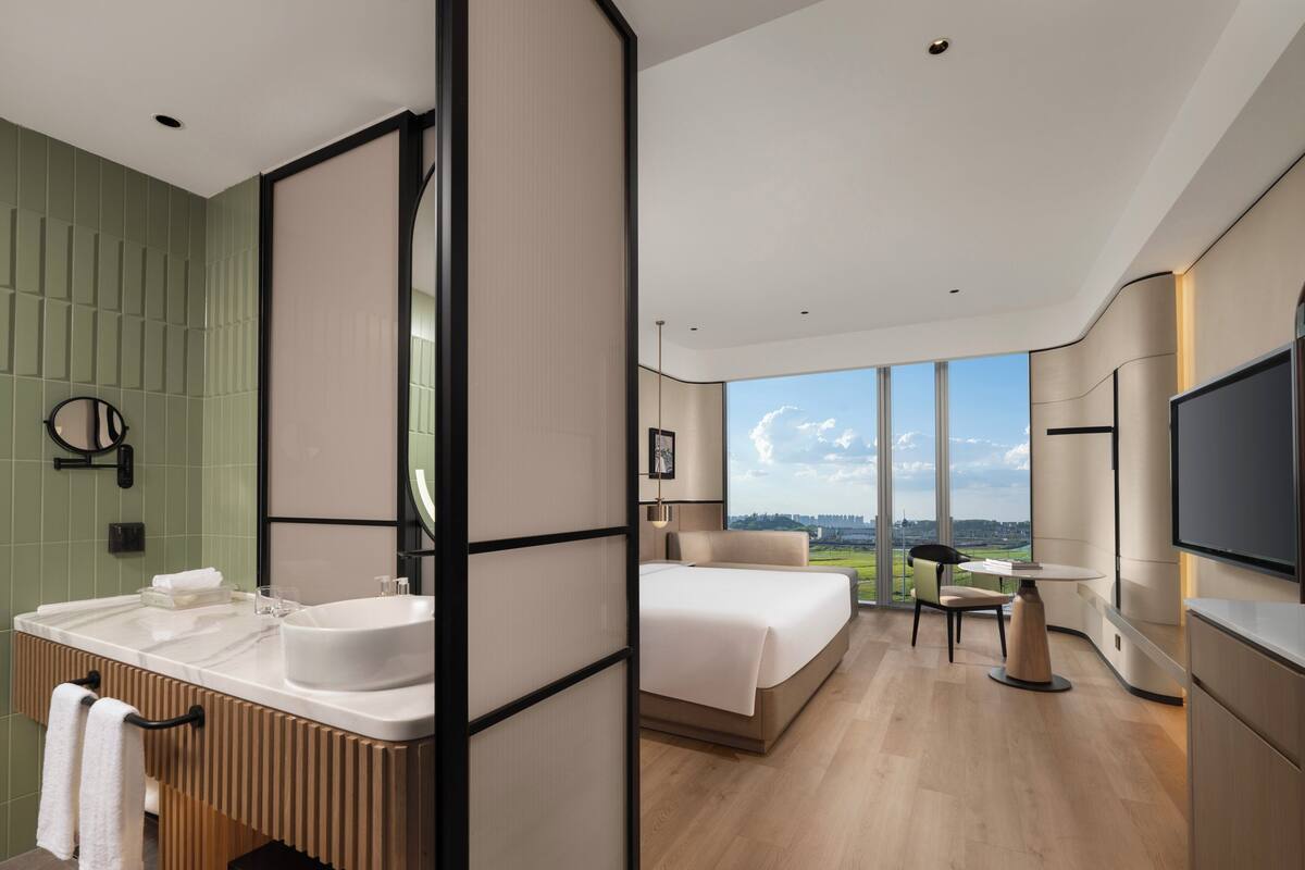 Ein modernes Hotelzimmer mit einem großen Bett, einem Schreibtisch, einem Fernseher und einem Badezimmer mit Waschbecken und Spiegel.