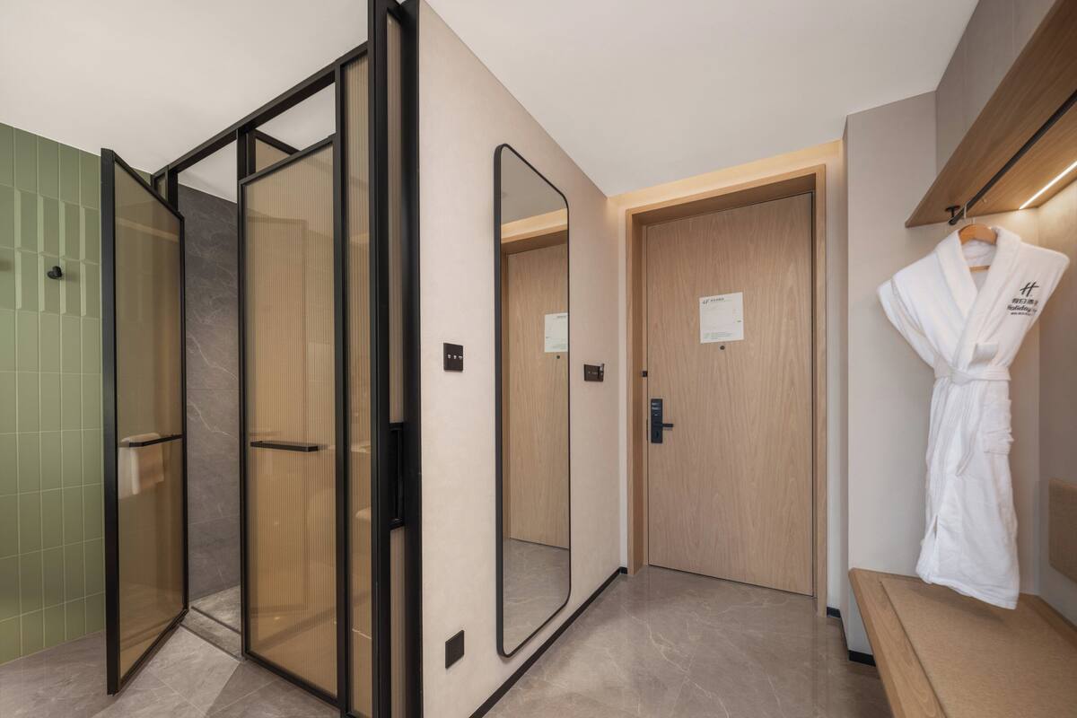 Ein Hotelbad mit Duschkabine aus Glas, einer Holztür und einem Bademantel, der an einem Haken hängt.
