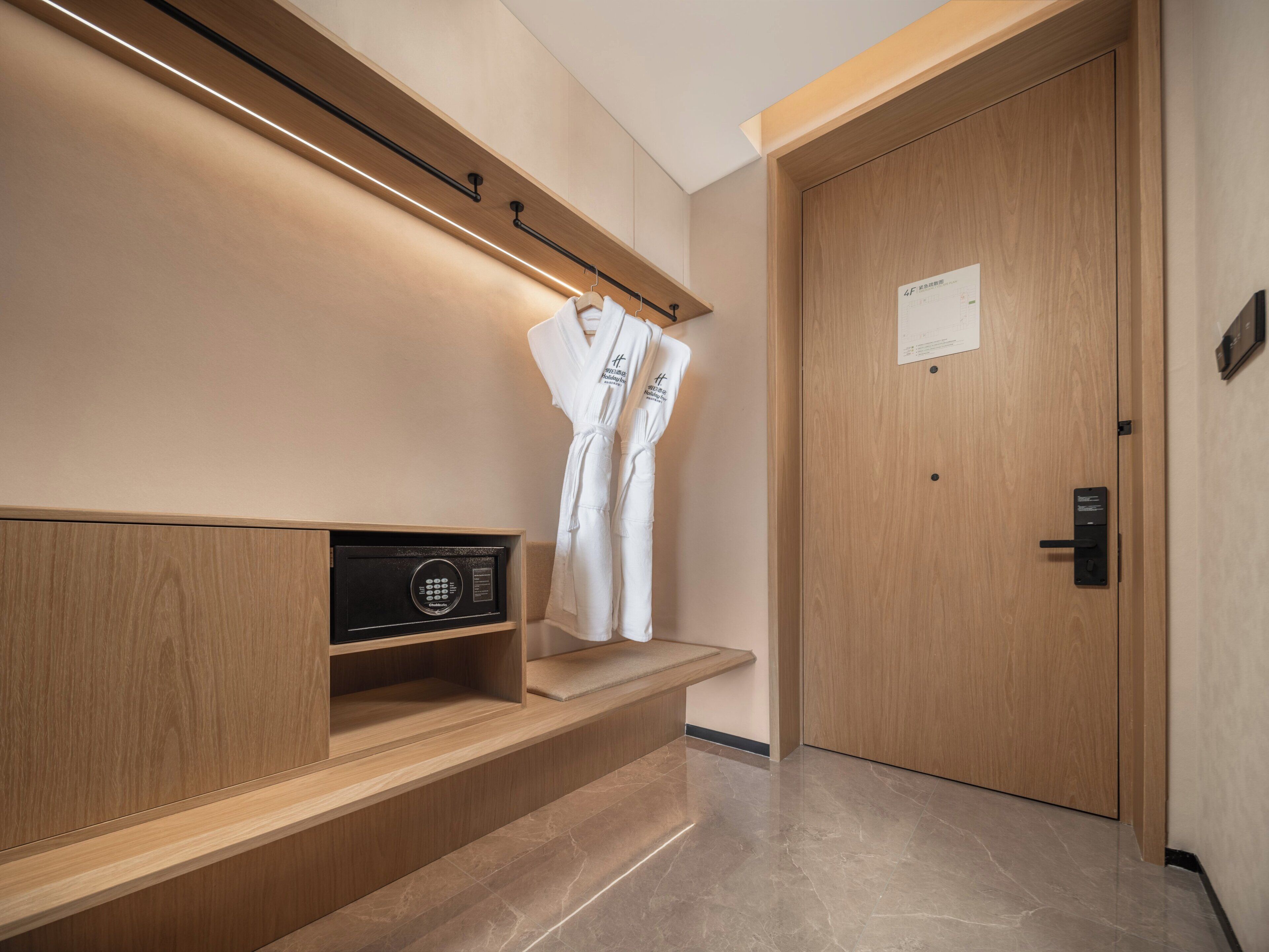 A modern hotel room with a wooden bench, a robe hanging on a hook, and a door with a sign.