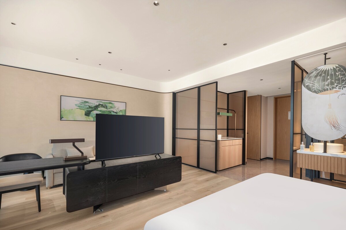 A modern hotel room with a large bed, a desk with a chair, a flat-screen TV, a wooden cabinet, and a bathroom with a mirror and sink.