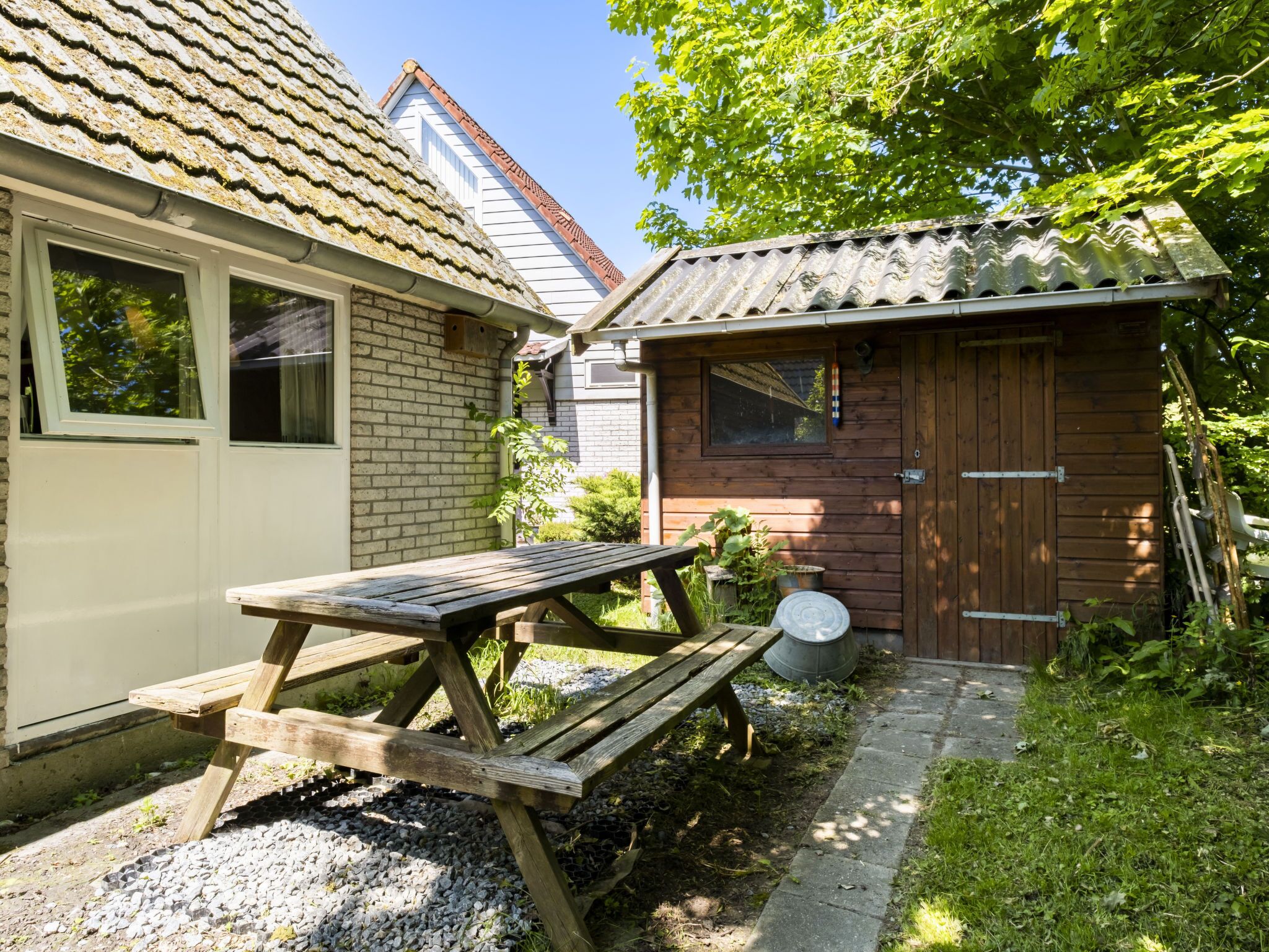 A wooden picnic table in a garden area with a brick house and a wooden shed in the background.