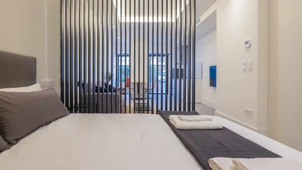 A modern hotel room with a large bed, a view through a vertical striped partition, and a small seating area visible through an open door. - Apartamento Presidente 1 (Sevilla)