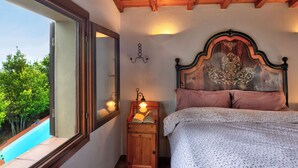 Une chambre avec un plafond en bois, un grand lit doté d’une tête de lit décorative, une table de chevet avec une lampe et une fenêtre donnant sur une piscine.