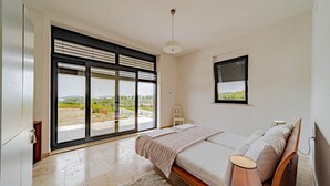 A bedroom with a large window, a bed, a bedside table, and a lamp.