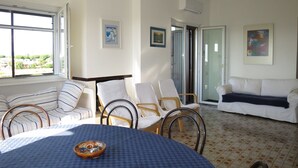 Een kamer met een ronde tafel, stoelen, een bank en een raam met uitzicht op groen.