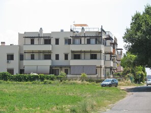 A multi-story residential building with balconies, a parking area, and a grassy field in front.