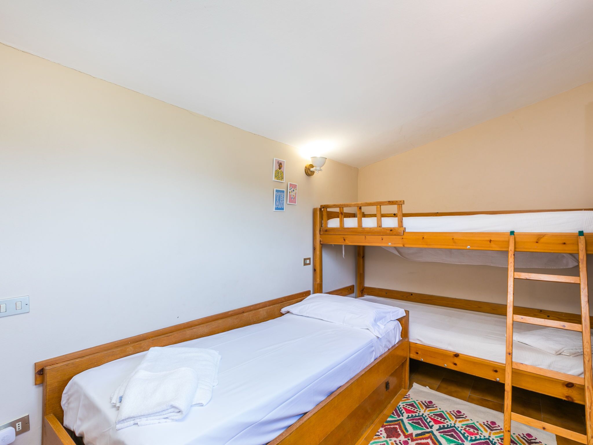 A bunk bed room with a single bed on the left and a bunk bed on the right. There is a wall-mounted light fixture above the single bed and a small wall decoration below it.
