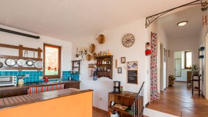 Una cocina con una isla de madera, una estufa y varios utensilios de cocina. Hay una ventana con cortinas, un reloj en la pared y una estantería con objetos de cocina.