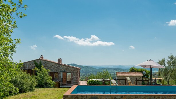 Una casa de piedra con piscina, rodeada de vegetación y colinas al fondo.