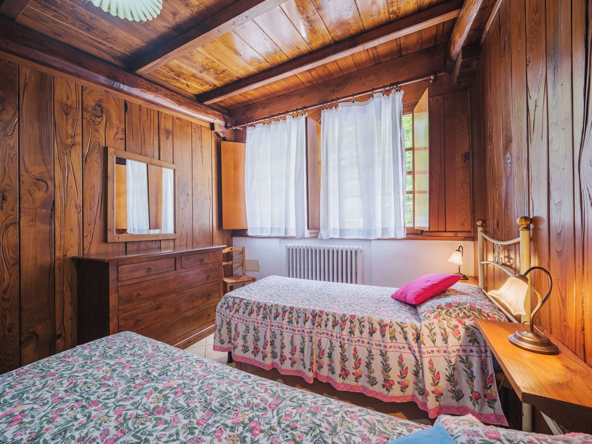 Een slaapkamer in een houten hutje met twee bedden, een dressoir, een spiegel en een raam met gordijnen.
