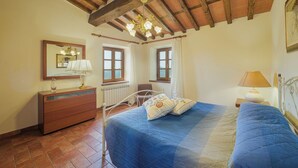 A bedroom with a wooden ceiling, two windows, a bed with blue bedding, a dresser, and a bedside table with a lamp. - Vacation home Sara by Interhome (Gello)