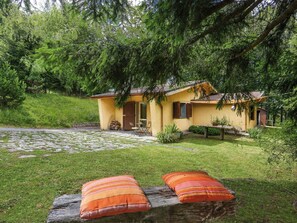 Una cabaña amarilla y acogedora con un patio de piedra, rodeada de exuberante vegetación y árboles.