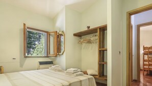 A bedroom with a bed, a window, a mirror, a shelf with clothes, and a wooden table. - Al Faggio (GLL120) by Interhome (Sillano)