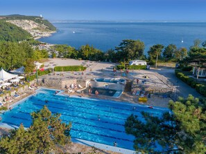 A large outdoor swimming pool with multiple lanes, surrounded by lounge chairs and umbrellas, with a view of the sea and hills in the background.
