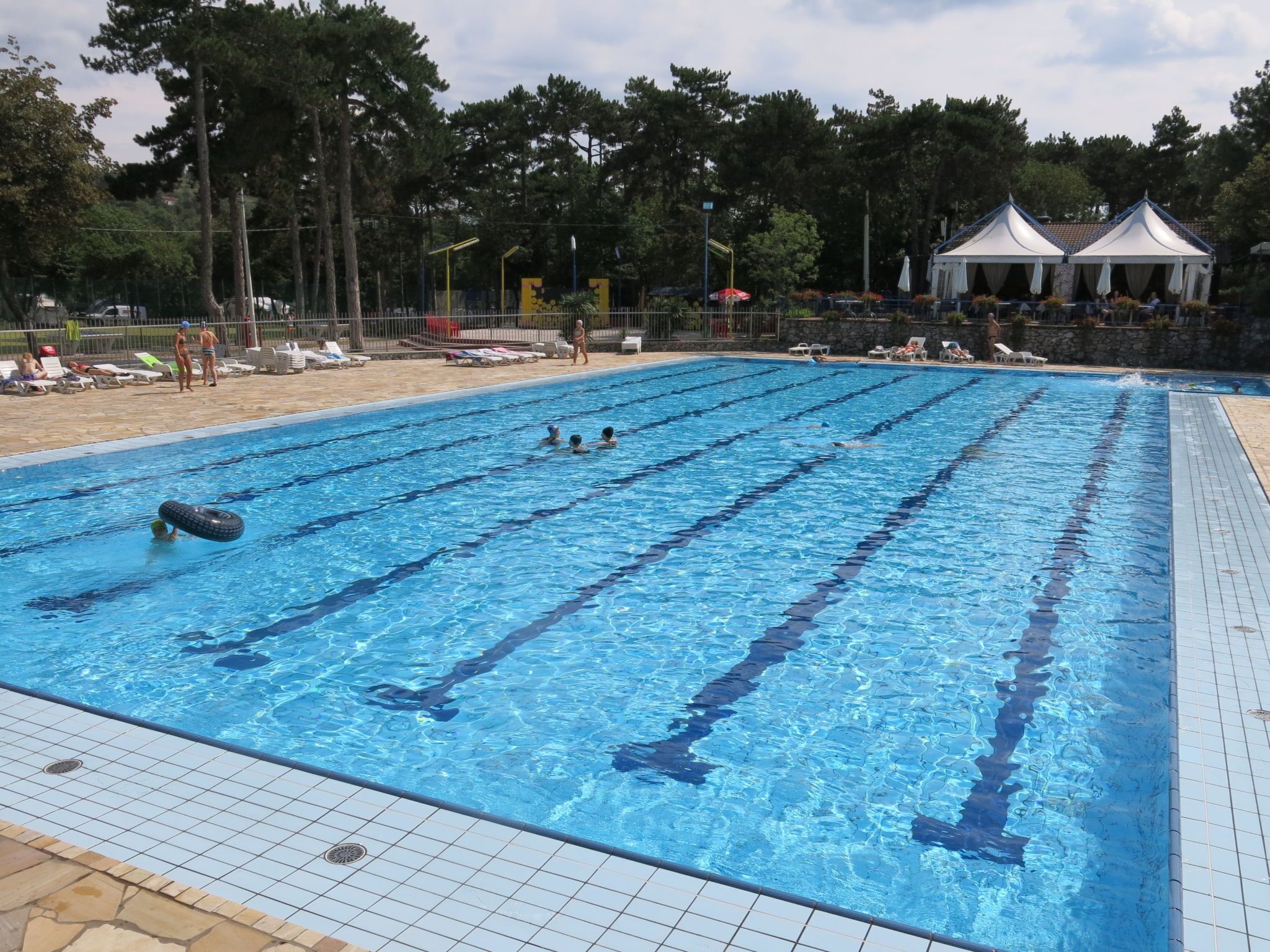 Una piscina con carriles delimitados, rodeada de tumbonas y una zona de estar cubierta.