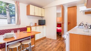 A compact kitchen with wooden cabinets, a dining table with stools, a television, and a window with curtains. - Camping Village Mare Pineta by Interhome (Sistiana-Duino-Aurisina)