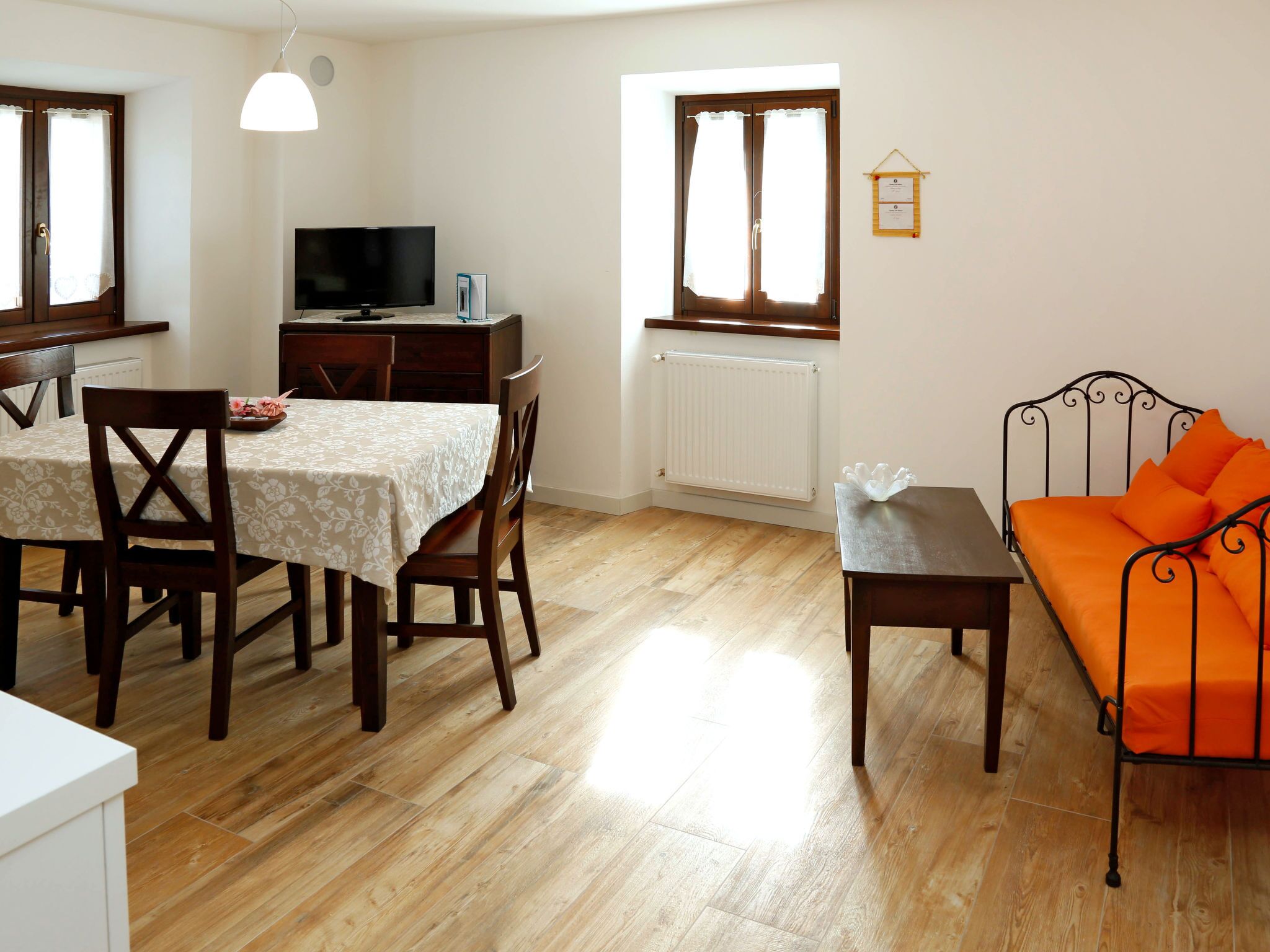A room with a dining table, chairs, a TV, a radiator, and a bed with orange bedding.