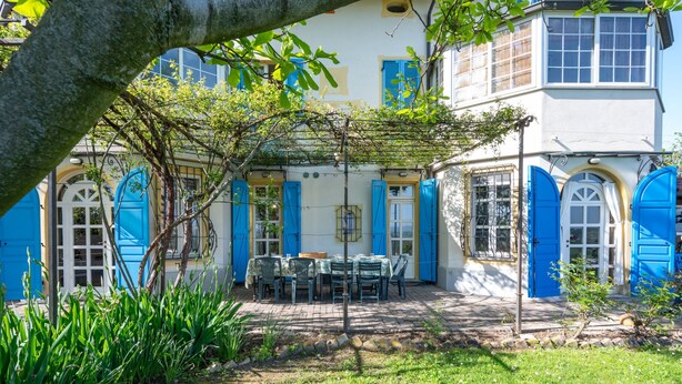 Un bâtiment à deux étages avec des volets et des portes bleues, un patio couvert avec une table et des chaises, et un jardin agrémenté de plantes et d’un arbre.
