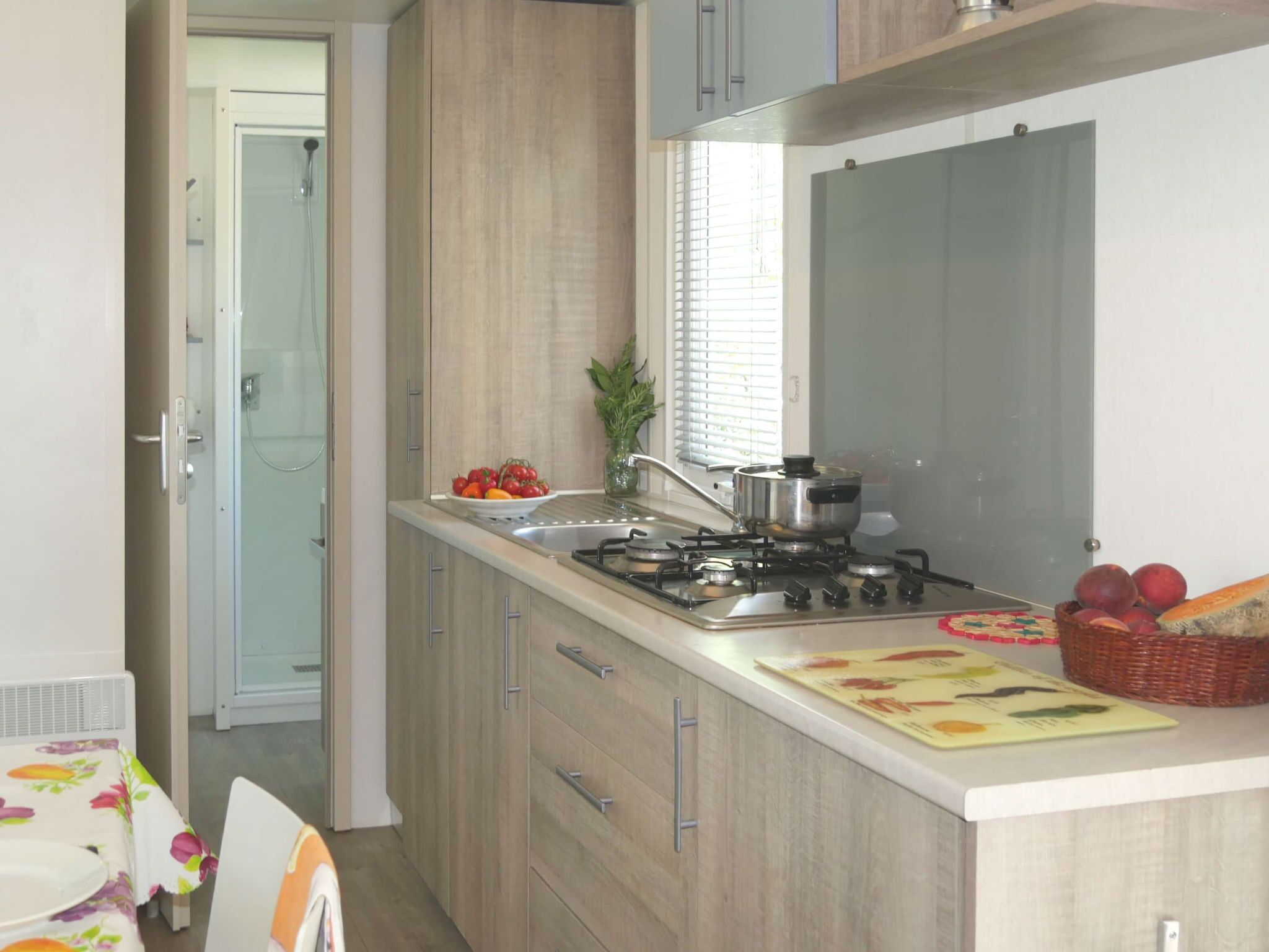 A modern kitchen with a gas stove, sink, and wooden cabinets. There is a window with blinds, a plant, and a basket of fruit on the counter.