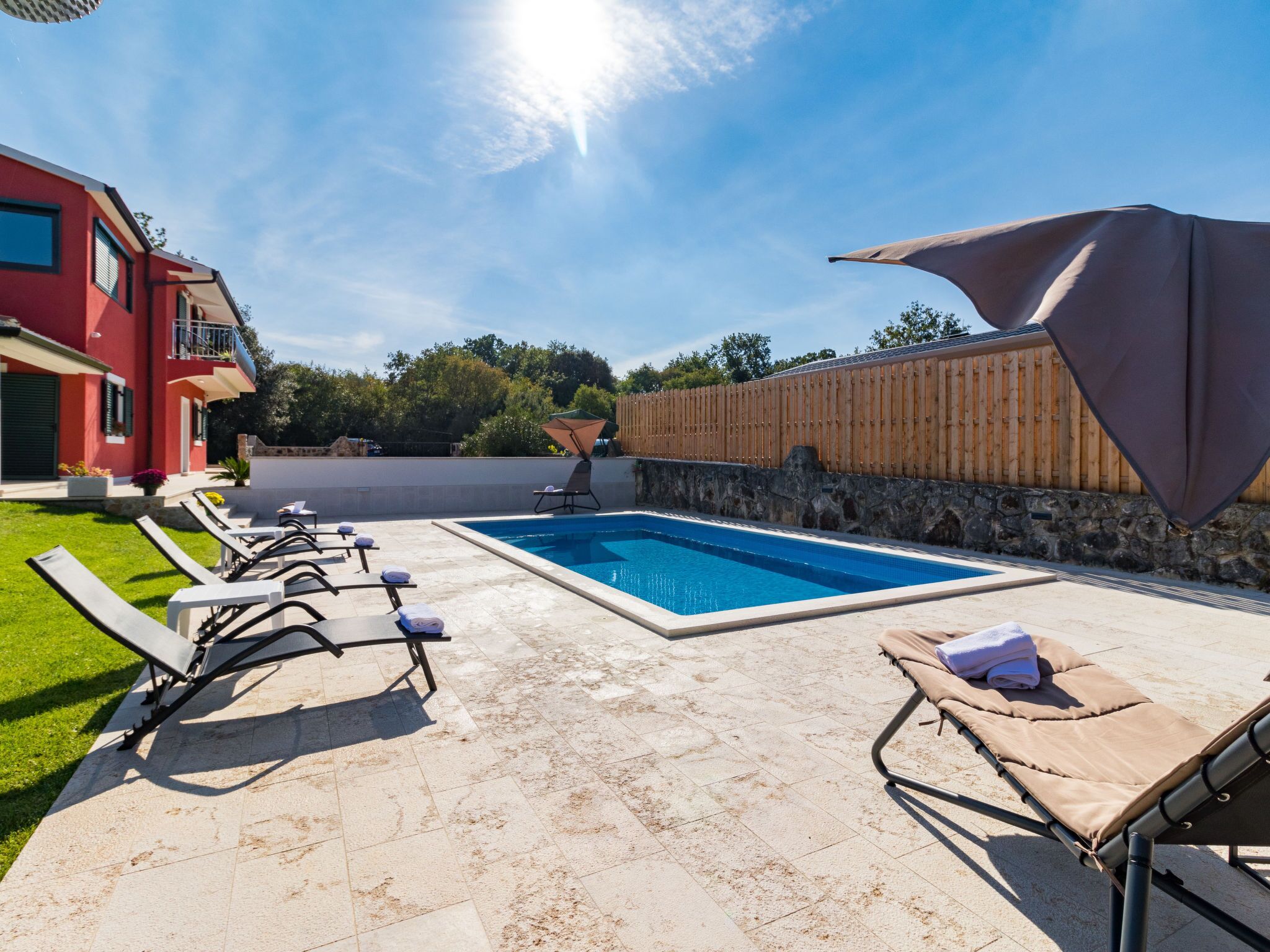 A swimming pool with lounge chairs, a wooden fence, and a red building in the background.