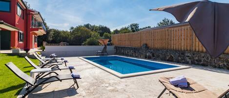 A swimming pool with lounge chairs, a wooden fence, and a red building in the background.