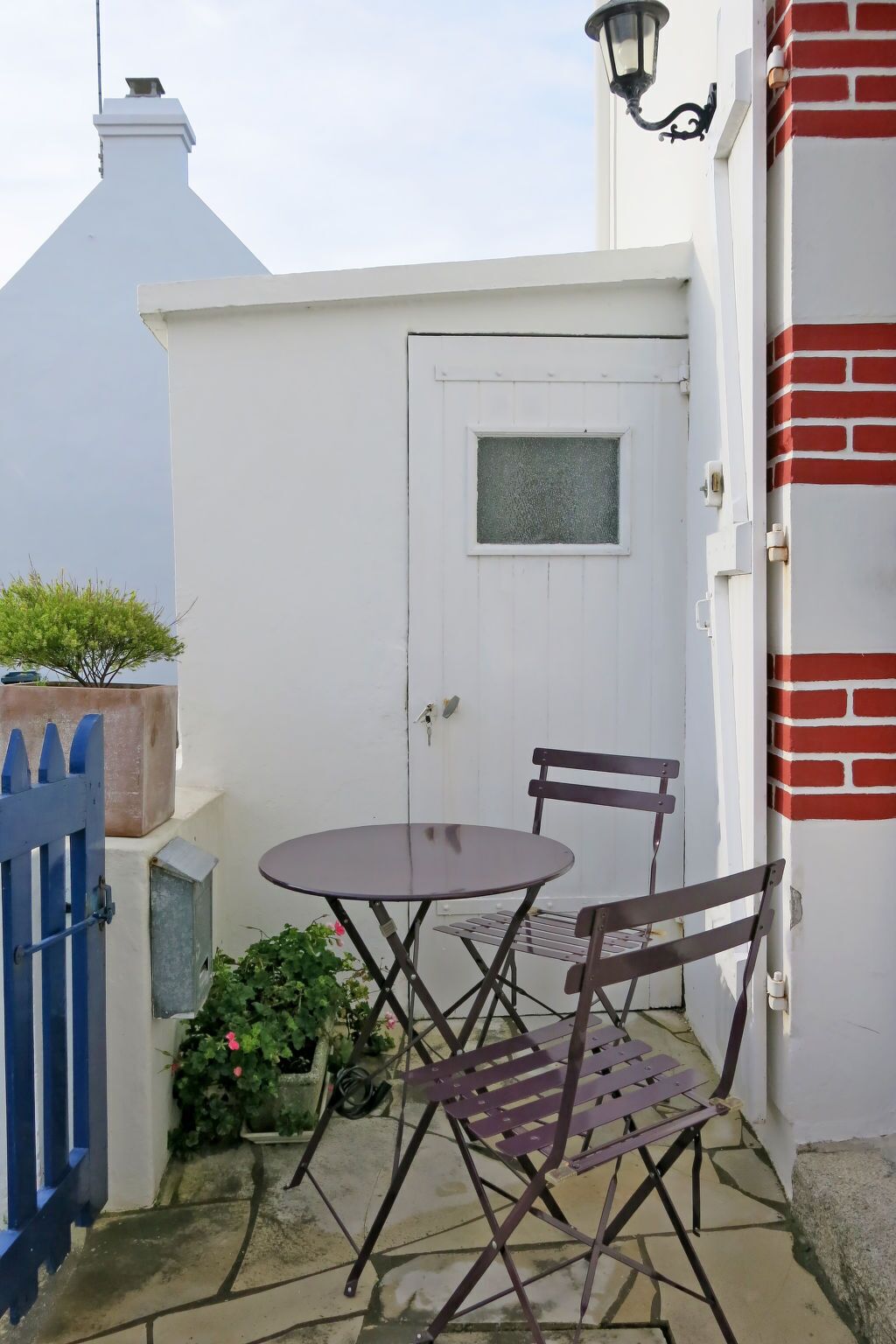 Eine kleine Terrasse im Freien mit einem runden Tisch und zwei Stühlen, einem blauen Tor und einer weißen Tür mit Fenster.