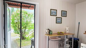 Una habitación con puerta corrediza de vidrio que da a un jardín, una mesa pequeña con una cesta de fruta, un estante de metal y dos cuadros en la pared.