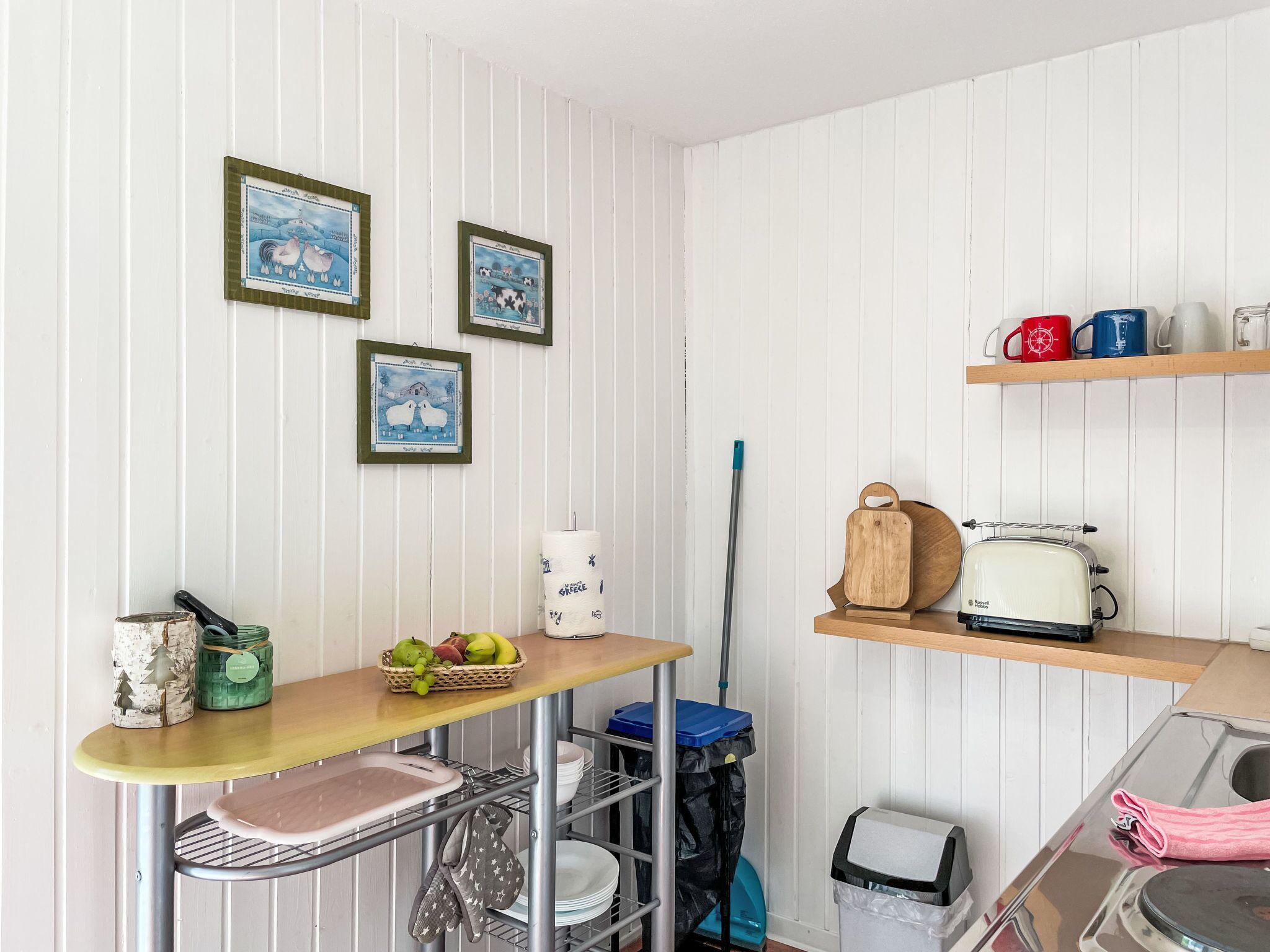 Une cuisine avec une table en bois, un grille-pain et un panier de fruits. Des tableaux encadrés ornent le mur, et une serpillière est appuyée contre le mur.
