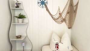 Una habitación con pared blanca, un mueble estante blanco curvo con una planta, una lámpara y una pequeña estatua. Hay una brújula decorativa y una red en la pared.