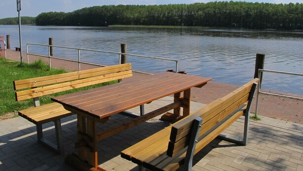 Un banco de madera y una mesa de picnic junto a un lago tranquilo, con un área de césped y una barandilla de metal.