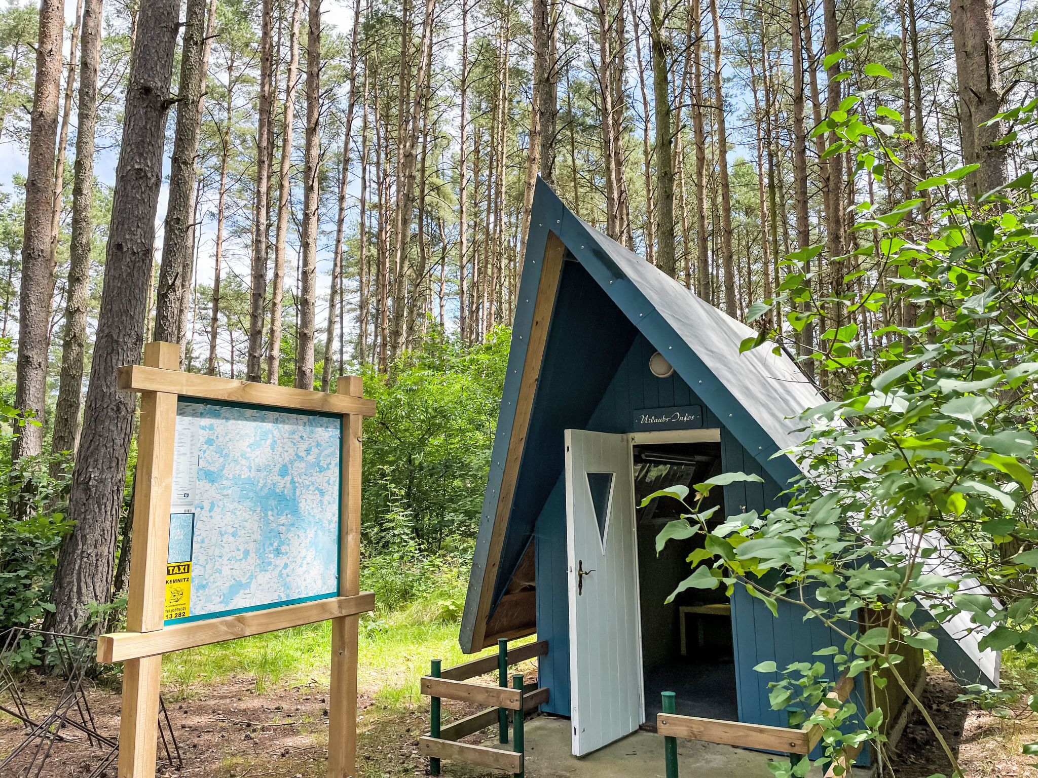 Une petite structure triangulaire en bois, dotée d’une porte et entourée d’une forêt.