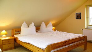 A single-bed room with a wooden frame, white bedding, two bedside lamps, a radiator, and a window with blinds.
