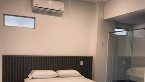 Un dormitorio moderno con cama, ventana, equipo de aire acondicionado y cabina de ducha de vidrio.