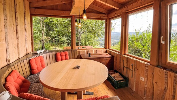 A wooden cabin with a round dining table, built-in bench seating, and large windows offering a view of the surrounding forest. - Mühlenmichelshäusle by Interhome (Hinterzarten)