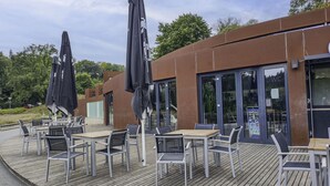 Ein Straßencafé mit Tischen und Stühlen, einer Holzterrasse und einem modernen Gebäude mit großen Fenstern.