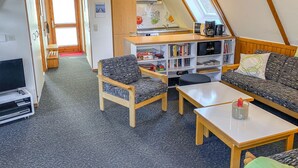 Ein gemütlicher, gut beleuchteter Raum mit Holzboden, einer Couch, einem Sessel, einem kleinen Tisch und einem Bücherregal voller Bücher.
