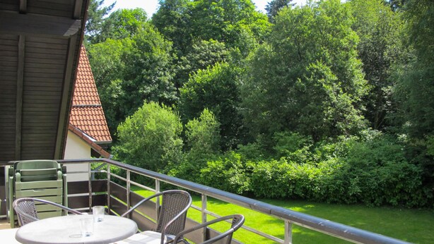 Un balcon avec une table et des chaises, donnant sur un jardin bien entretenu et des arbres.