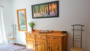 Une chambre avec une commode en bois, un lit recouvert d’une couvre-lit à motifs, une plante en pot, un tableau encadré et une échelle.
