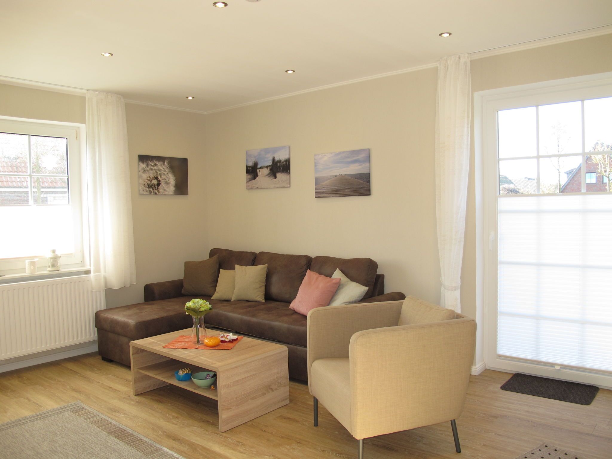 A modern living room with a brown leather sofa, a beige armchair, and a wooden coffee table. There are three framed pictures on the wall, and the room has large windows with white curtains.
