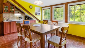 A dining area with a wooden table and chairs, a kitchen with yellow walls and wooden beams, and large windows offering a view of greenery.