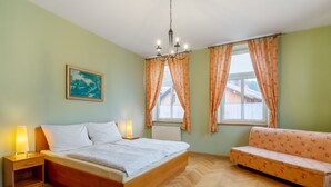 A bedroom with a wooden bed, two bedside lamps, a window with curtains, a sofa, and a painting on the wall. - Base Camp 1 by Interhome (Desná v Jizerských horách)