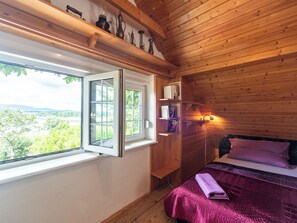A wooden cabin bedroom with a large window, a bed with a purple blanket, and a shelf with books and a lamp.