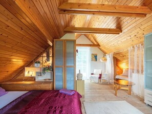 Ein gemütliches Schlafzimmer unter dem Dach mit geneigter Decke, Holzbalken, einem Bett mit violetter Decke, einem kleinen Tisch und einem Stuhl.