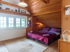 Une chambre confortable avec un plafond en bois, un lit recouvert d’une couverture violette, une lampe de chevet et une fenêtre donnant sur un beau paysage.