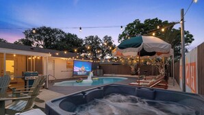 A backyard with a hot tub, a swimming pool, outdoor seating, and a TV screen displaying a movie.