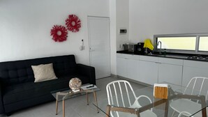 Uma sala de estar moderna com um sofá preto, uma mesa de centro de vidro e cadeiras brancas. Uma cozinha com armários brancos e uma janela é visível ao fundo.
